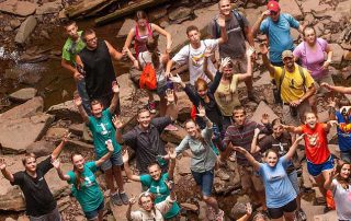 a bunch of wayumi participants on the edge of a rocky stream bed waving something up above them